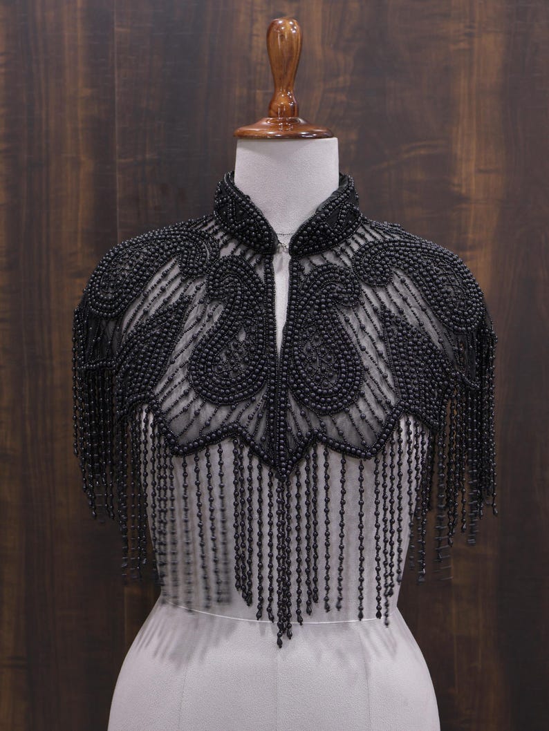 May include: A black beaded capelet with a high collar and intricate paisley-like designs. The capelet features long beaded fringe that hangs down from the shoulders. The garment is displayed on a mannequin against a dark wood background.