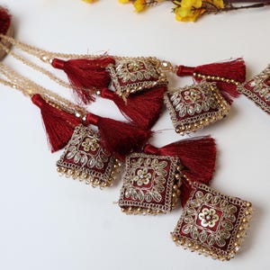 May include: Maroon and gold decorative tassels with square, embroidered embellishments. Each square is adorned with gold thread and small beads, and is attached to a gold beaded chain. The tassels are a deep maroon color.