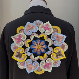 Può includere: Giacca nera con un colorato motivo floreale ricamato sul retro. Il ricamo presenta un motivo simmetrico con accenti rosa, blu, gialli e marroni. La giacca è esposta su un manichino.