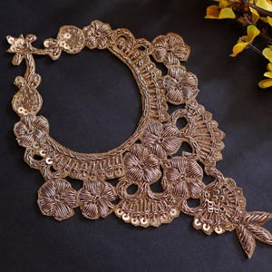 May include: Gold-coloured embroidered trim with a floral design and sequins. The trim is shaped like a teardrop and has a circular opening in the centre.