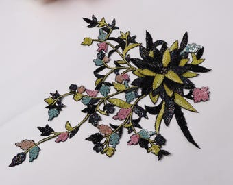 May include: Embroidered floral applique featuring a cluster of black and yellow flowers with a trailing vine. The vine has green, blue, and pink leaves and small flowers. This decorative applique is ideal for embellishing clothing or accessories.