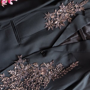 Puede incluir: Una chaqueta de color verde oscuro con dos adornos florales decorativos hechos de lentejuelas y cuentas.