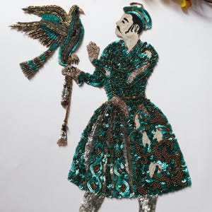 May include: An embroidered applique depicting a figure in a green and gold outfit, holding a bird. The figure's attire is embellished with sequins and beads. The bird has teal and gold accents. The applique is likely for decorative purposes.