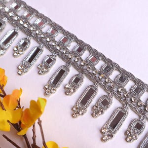May include: Silver trim with a fringe of small rectangular mirrors and rhinestones. The trim is on a white background.