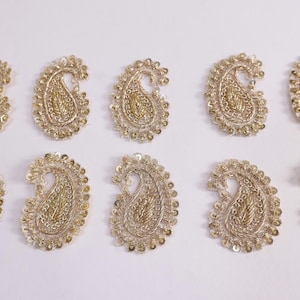 May include: A collection of ten gold paisley appliques, each with detailed embroidery and sequin embellishments. The appliques are arranged on a white surface. The intricate design and gold colour create a decorative and ornate appearance.