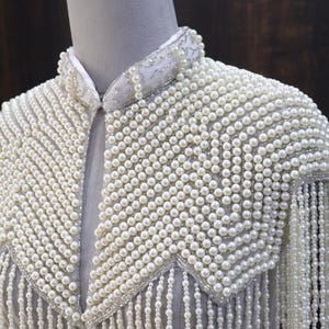 May include: A white, pearl-covered garment with a collar and shoulder embellishments. The piece features a dense arrangement of small, round pearls, with longer strands of pearls hanging from the shoulders, creating a fringed effect. The garment is displayed on a mannequin.