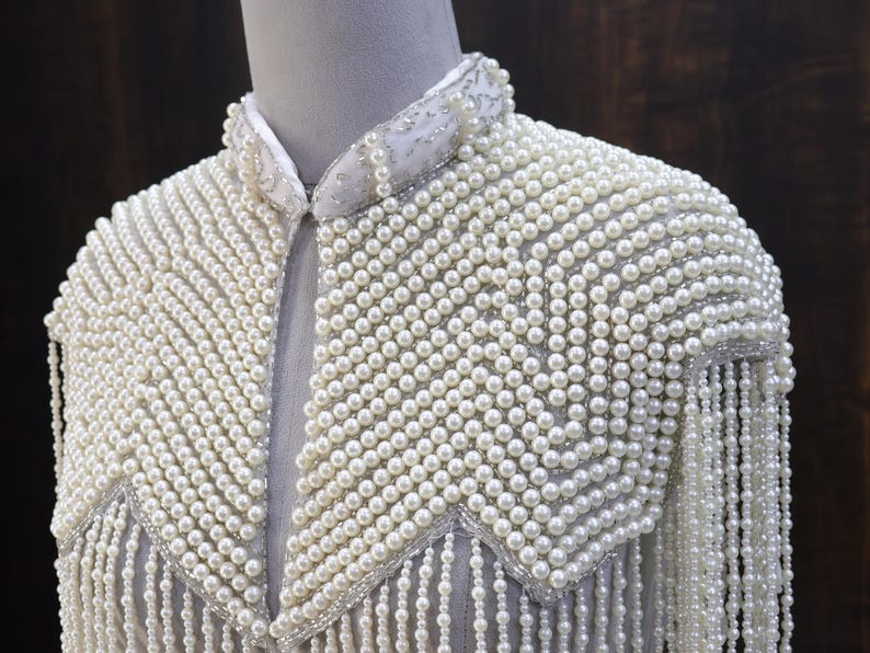 May include: A white, pearl-covered garment with a collar and shoulder embellishments. The piece features a dense arrangement of small, round pearls, with longer strands of pearls hanging from the shoulders, creating a fringed effect. The garment is displayed on a mannequin.