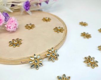 May include: Gold-toned flower-shaped embellishments with clear crystal accents. The floral designs are scattered on a beige surface and a wooden embroidery hoop. Decorative floral elements are in the background.