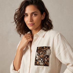 May include: A beige button-down shirt featuring a rectangular chest pocket adorned with a detailed tiger head patch. The patch is crafted with black, gold, and white sequins and beads, depicting a tiger face surrounded by floral accents.