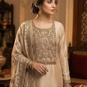 May include: A cream-colored, long-sleeved dress with intricate gold embroidery on the bodice and sleeves. The dress is paired with a matching shawl, and the model is wearing gold bangles. The overall style is elegant and formal.