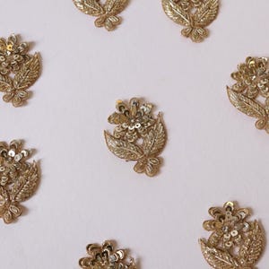 May include: A collection of eight gold-toned floral appliques arranged in a circular pattern. Each individual piece features a central flower with sequin detailing and two textured leaves at the base, crafted with metallic thread embroidery.
