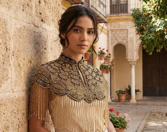 May include: A cream-colored sleeveless dress featuring a sheer black overlay capelet adorned with intricate gold embroidery and long, dangling gold beaded fringe that extends over the shoulders and chest, creating a layered and textured appearance.