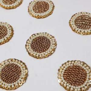 Puede incluir: Primer plano de varios adornos circulares decorativos. Cada uno tiene un centro dorado tejido, rodeado por un anillo de pequeñas perlas blancas y lentejuelas doradas. Los adornos están dispersos sobre una superficie blanca.