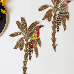 May include: Two gold embroidered patches featuring a bird perched on a palm tree. The birds are decorated with colourful beads and sequins. The patches are perfect for adding a touch of whimsy to clothing or accessories.