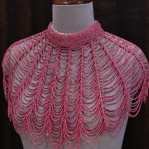 May include: A pink beaded collar necklace with a high neckline and a cascading design. The necklace is made of small, round beads in shades of pink and gold, creating a textured and elegant look. The necklace is displayed on a mannequin.