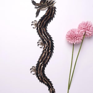May include: A detailed, beaded dragon applique in black, brown, and gold. The dragon has a long, serpentine body with intricate detailing, claws, and a fierce expression. Two pink flowers with green stems are to the right.