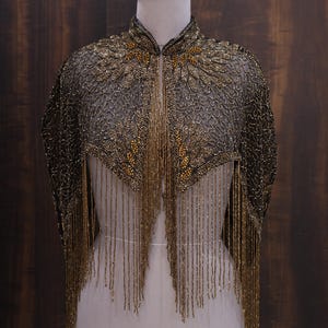 May include: A gold and black beaded cape with a high collar and long fringe. The cape is displayed on a mannequin against a dark wood backdrop. The intricate beadwork creates a floral pattern.