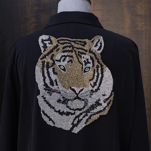 Peut inclure: Chemise noire avec une tête de tigre brodée dans le dos. Le tigre est orné de perles dorées, argentées et noires, avec des yeux verts et un nez rose. La chemise est présentée sur un mannequin.