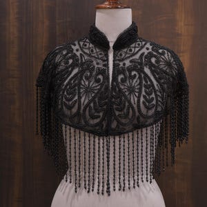 May include: Black beaded capelet with a high collar and intricate floral patterns. The capelet features a zipper closure and long beaded fringe. The garment is displayed on a mannequin against a dark wood background.