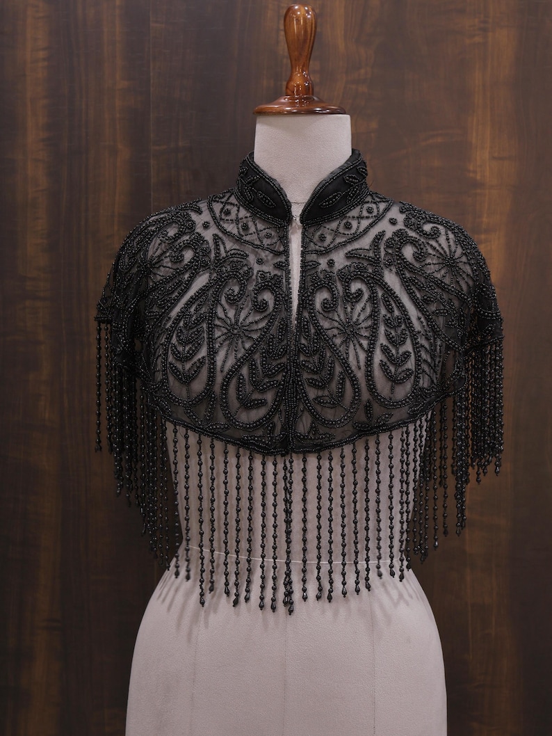 May include: Black beaded capelet with a high collar and intricate floral patterns. The capelet features a zipper closure and long beaded fringe. The garment is displayed on a mannequin against a dark wood background.