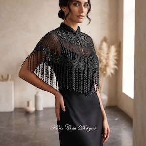 May include: A black sleeveless dress paired with a sheer black beaded capelet featuring a high collar, intricate embroidery, and long fringe detailing. The garment displays the text Kora Casa Designs in white script near the waistline area.