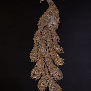 May include: An ornate gold-coloured peacock design, featuring detailed beadwork and a red jewel eye. The peacock's tail feathers are fanned out, creating an elegant appearance. The design is set against a black background.