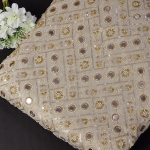 Könnte beinhalten: Cremefarbenes Zierkissen mit einem geometrischen Muster aus goldener Stickerei, Pailletten und kleinen Spiegelakzenten. Ein Strauß weißer Blumen liegt in der Nähe des Kissens. Das Kissen ist quadratisch und scheint aus einem strukturierten Stoff zu bestehen.