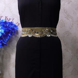 May include: A gold and silver embellished belt with a floral design is worn over a black dress. The belt features a repeating pattern of gold sequins and silver mirrored accents. The dress is sleeveless and has a button-down front.