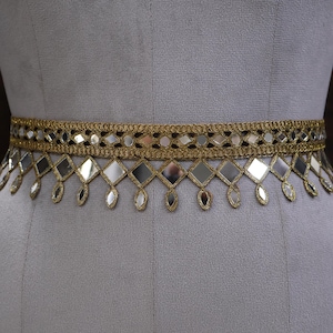 Peut inclure: Une ceinture dorée ornée de motifs en forme de diamants et de gouttes miroitantes. La ceinture est portée autour de la taille d'un mannequin, mettant en valeur son design décoratif. Les détails complexes et les surfaces réfléchissantes créent un look glamour.