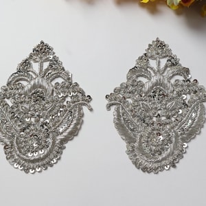 Puede incluir: Dos apliques bordados plateados con un diseño floral simétrico. Cada aplique presenta intrincados abalorios y lentejuelas, creando un efecto brillante. Los apliques son ideales para embellecer prendas o accesorios.
