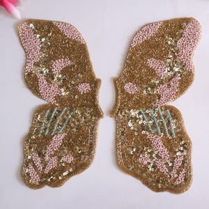 May include: A pair of gold butterfly-shaped fabric appliques with pink and blue sequins. The appliques are perfect for adding a touch of sparkle to clothing, bags, or home decor.