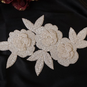 May include: Ivory beaded floral applique featuring three flower blooms and three leaves. The flowers and leaves are made of small, shiny beads. The applique is set against a black background.