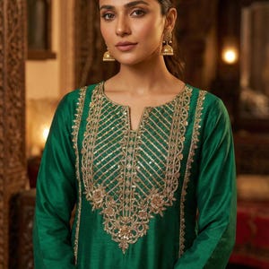 May include: A woman wearing a long-sleeved, emerald green tunic with intricate gold embroidery around the neckline and cuffs. The tunic has a V-neck design and is adorned with sequins. The woman is also wearing gold earrings.