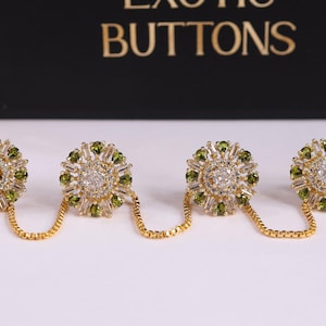 May include: Four gold-toned buttons with a floral design, each adorned with clear and green gemstones. The buttons are connected in pairs by a delicate gold chain. The text "EXOTIC BUTTONS" is visible in the background.