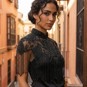 May include: A black beaded top with a high collar and fringe detailing. The top is made of a sheer fabric and is adorned with intricate beadwork. The model is standing outside in front of a building with an orange facade.