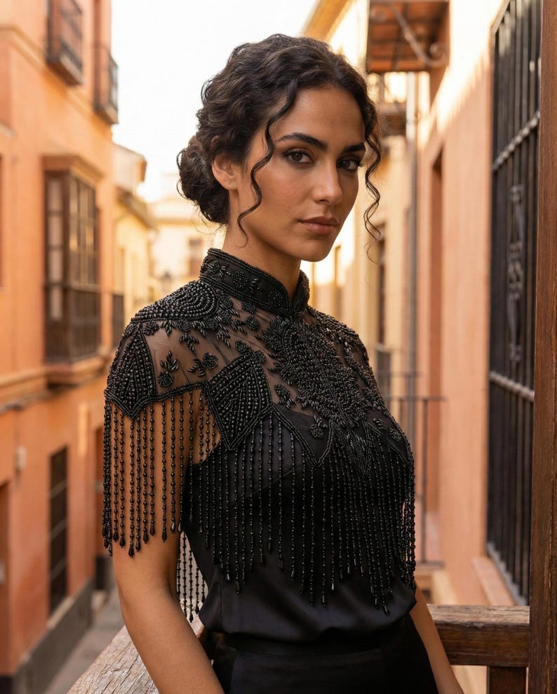 May include: Black beaded top with a high collar and fringe detailing. The top features intricate beadwork on the shoulders and chest, with long beaded fringe hanging down. The garment is worn over a black top.