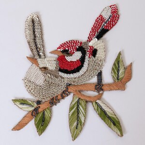 Può includere: Una toppa decorativa raffigurante due uccelli appollaiati su un ramo. Gli uccelli sono realizzati con paillettes bianche, rosse e nere. Il ramo è marrone con foglie verdi accentuate da perline scure. Il design presenta ricami dettagliati.