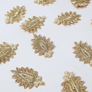 Puede incluir: Adornos dorados en forma de hoja con intrincados bordados y pequeños acentos de perlas blancas. Estos elementos decorativos están dispuestos sobre una superficie blanca, mostrando su diseño detallado.