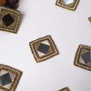 Puede incluir: Seis adornos decorativos de forma de diamante, de color dorado, con un pequeño espejo en el centro. Cada adorno está rodeado por un borde de cuentas.