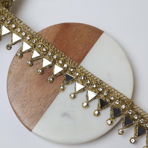 May include: Gold-colored trim with mirrored triangle accents and clear crystal-like embellishments. The trim is displayed on a two-tone, round surface of wood and white marble. The trim has a decorative, ornate design.