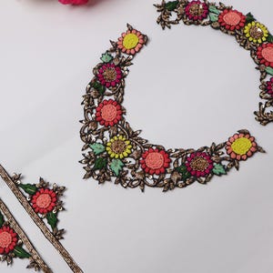 May include: Embroidered floral appliques in a semi-circular design, featuring pink, yellow, and coral flowers with green leaves and gold accents. Includes two strips of matching trim. Ideal for embellishing garments or crafts.