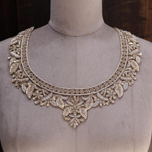 Peut inclure: Un col brodé argenté et doré avec un motif floral et de feuilles. Le col est présenté sur un mannequin gris. La broderie présente des paillettes et des détails complexes, créant un décolleté décoratif.