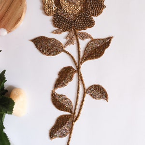 Puede incluir: Aplicación floral decorativa con una gran rosa dorada de lentejuelas y un centro de perlas. El tallo y las hojas están detallados con cuentas doradas. Ideal para añadir un toque elegante a prendas o manualidades. Medidas en centímetros.