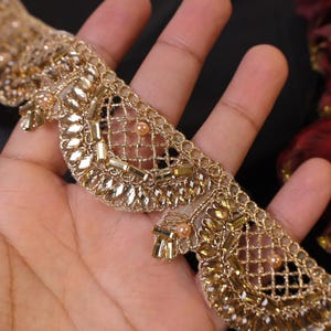 May include: Close-up of a gold-colored decorative trim. The trim features a net-like design with small, rectangular and oval-shaped embellishments. Small, round beads are also incorporated into the design. The trim is held in a hand.