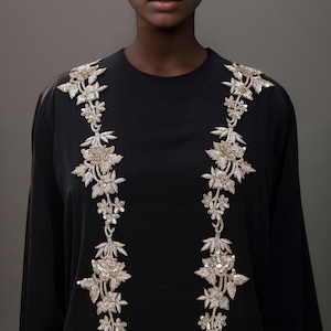 Puede incluir: Una camisa negra de manga larga con cuello en V y dos filas verticales de bordados florales dorados y plateados.