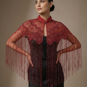 May include: A burgundy beaded cape with a high collar and detailed floral patterns. The cape has long fringe detailing on the sleeves and front, creating a flowing, elegant look. The cape is worn over a black dress.