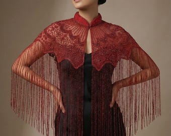 May include: A burgundy beaded cape with a high collar and detailed floral patterns. The cape has long fringe detailing on the sleeves and front, creating a flowing, elegant look. The cape is worn over a black dress.