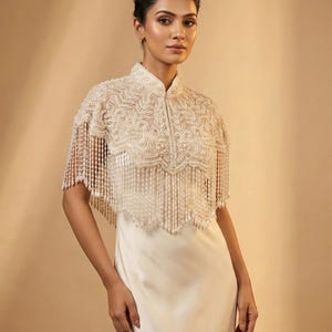 May include: Ivory-colored beaded capelet with intricate floral embroidery and fringe detailing. The capelet features a high collar and is worn over a white dress, creating an elegant and sophisticated look. The garment is suitable for special occasions.