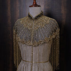 Op de afbeelding: Een gouden kralen cape met een hoge kraag en franjedetails. De cape heeft ingewikkeld kralenwerk rond de halslijn en schouders, met lange strengen kralen die naar beneden vallen. Een dunne gouden riem accentueert de taille.
