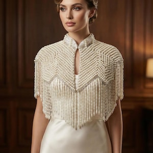 May include: Ivory beaded bridal capelet with fringe. The capelet features a high collar and a geometric pattern of small beads. The fringe hangs from the shoulders, adding movement and elegance to the design. The capelet is worn over a white satin dress.
